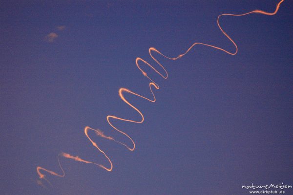 Kondensstreifen von Flugzeug im rötlichen Morgenlicht, vom Wind zu stark gewundener Linie verformt, Göttingen, Deutschland