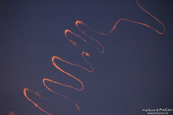 Kondensstreifen von Flugzeug im rötlichen Morgenlicht, vom Wind zu stark gewundener Linie verformt, Göttingen, Deutschland